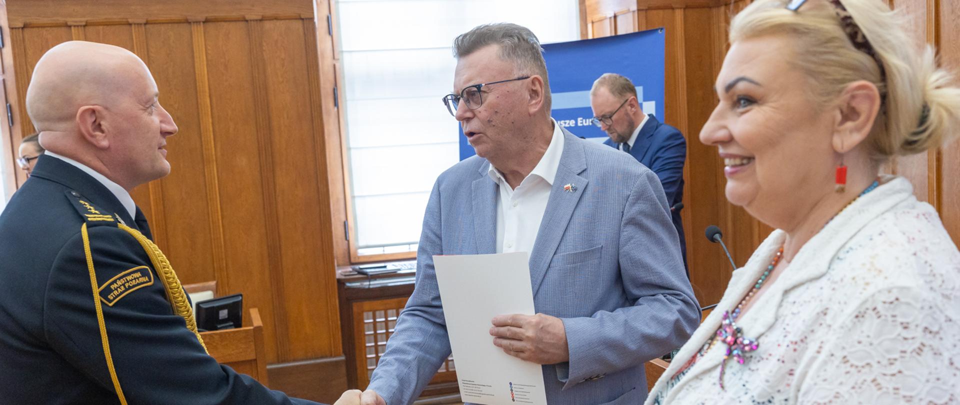 Mężczyzna w szarym garniturze trzyma dokument i wita się uściskiem dłoni ze strażakiem w mundurze galowym Państwowej Straży Pożarnej. Obok nich stoi uśmiechnięta kobieta w białej, koronkowej marynarce, która przygląda się powitaniu.