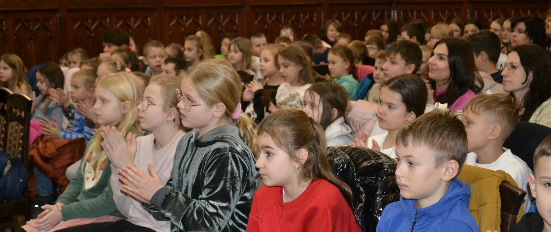 Zdjęcie przedstawia grupę dzieci i dorosłych siedzących w eleganckiej sali z drewnianą boazerią. Część dzieci klaszcze, a inne skupiają się na wydarzeniu poza kadrem. 