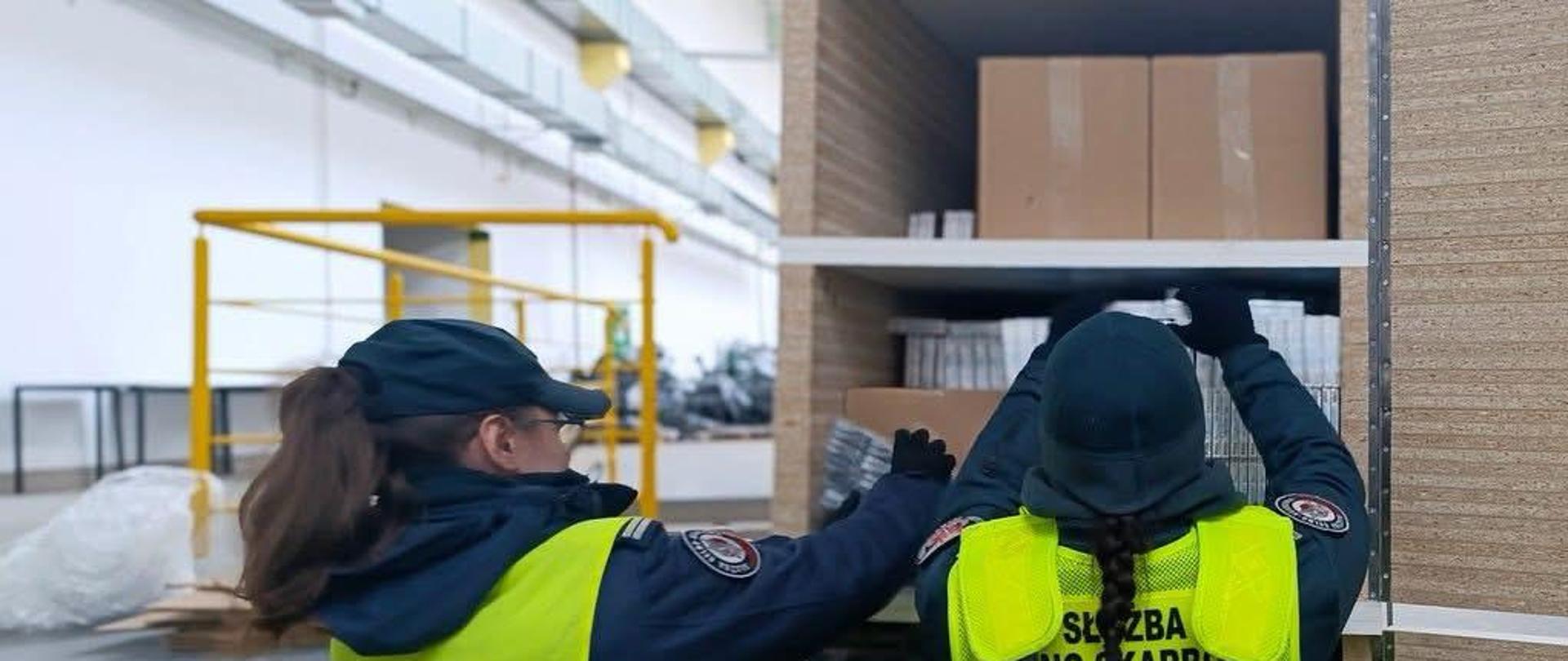 Dwie osoby w kamizelkach z napisem "Służba-Celno-Skarbowa" w tle widać elementy hali magazynowej oraz żółtą barierkę