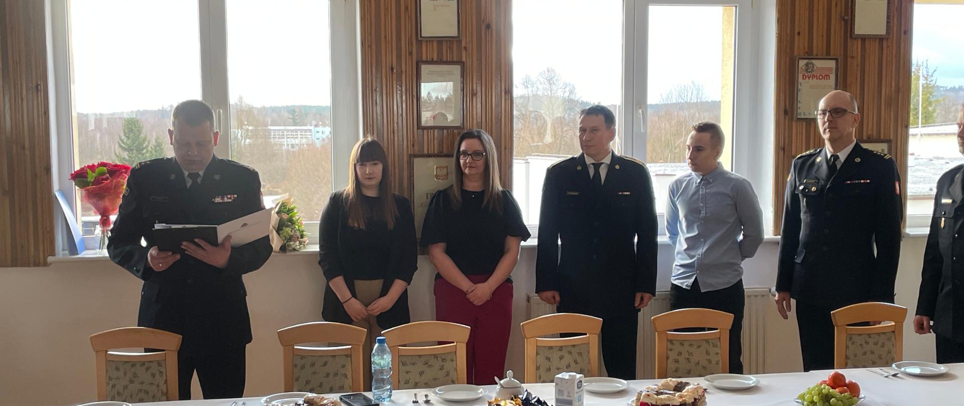 Osoby w szeregu, jedna osoba ubrana w ciemny w mundur trzyma w ręku białą teczkę. 