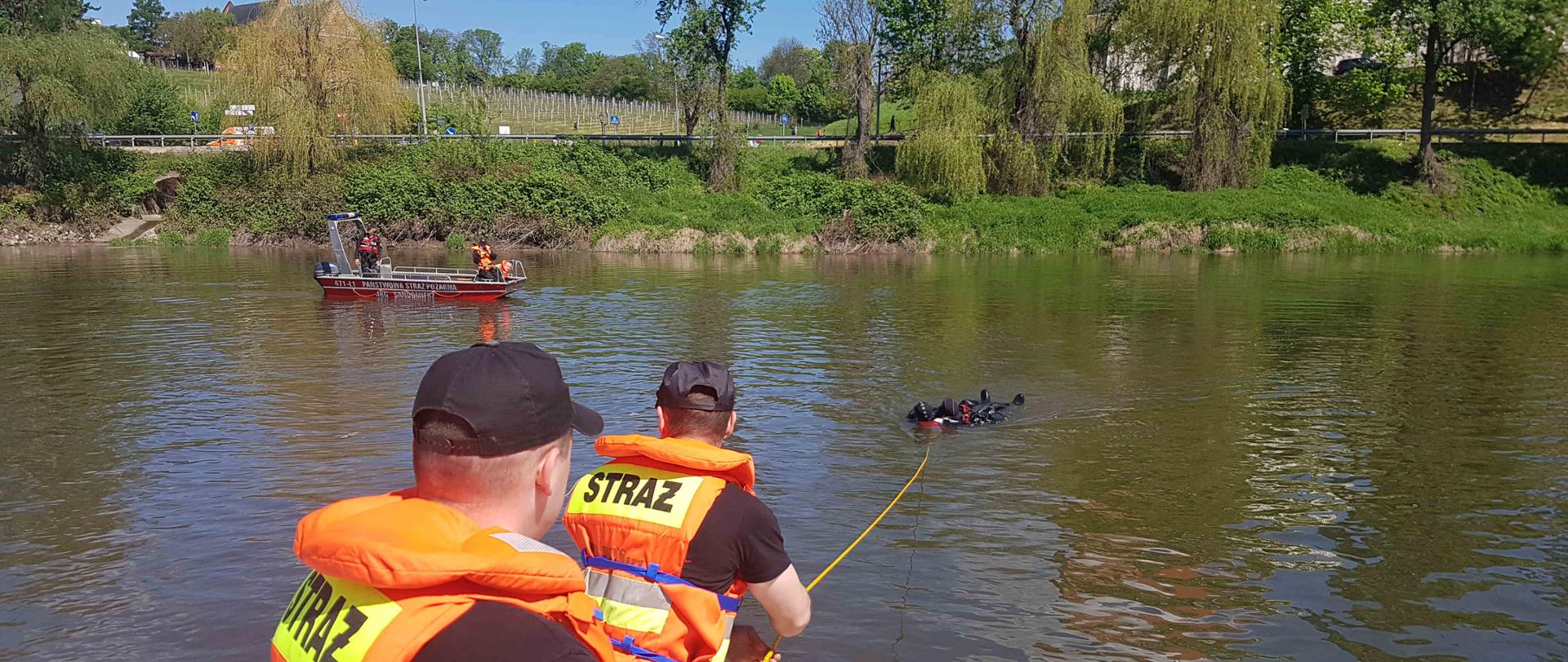 zdjęcie zrobione w dzień. Zdjęcie przedstawia zbiornik wodny w którym jest jeden strażak wodzie i woła o pomoc. Dwóch strażaków z brzegu przy pomocy linki i jednego strażaka w wodzie wyciągają poszkodowanego. W oddali widać płynąca łódkę, na której znajduje się trzech strażaków. 