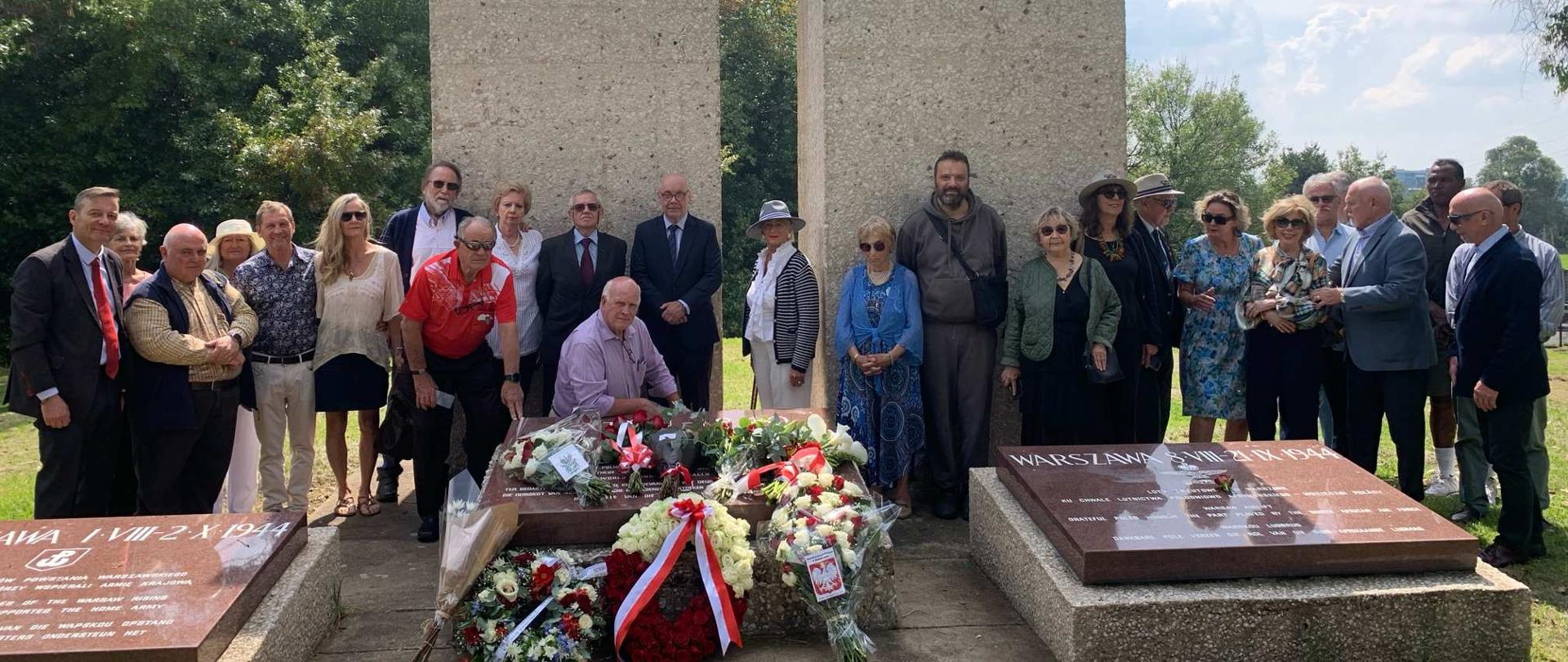 Grupa 25 osób - uczestników obchodów Dnia Pamięci Ofiar Zbrodni Katyńskiej pozuje przed pomnikiem Katyńskich w Johannesburgu; na pierwszym planie 3 tablice pamięci oraz biało-czerwone wieńce i wiązanki