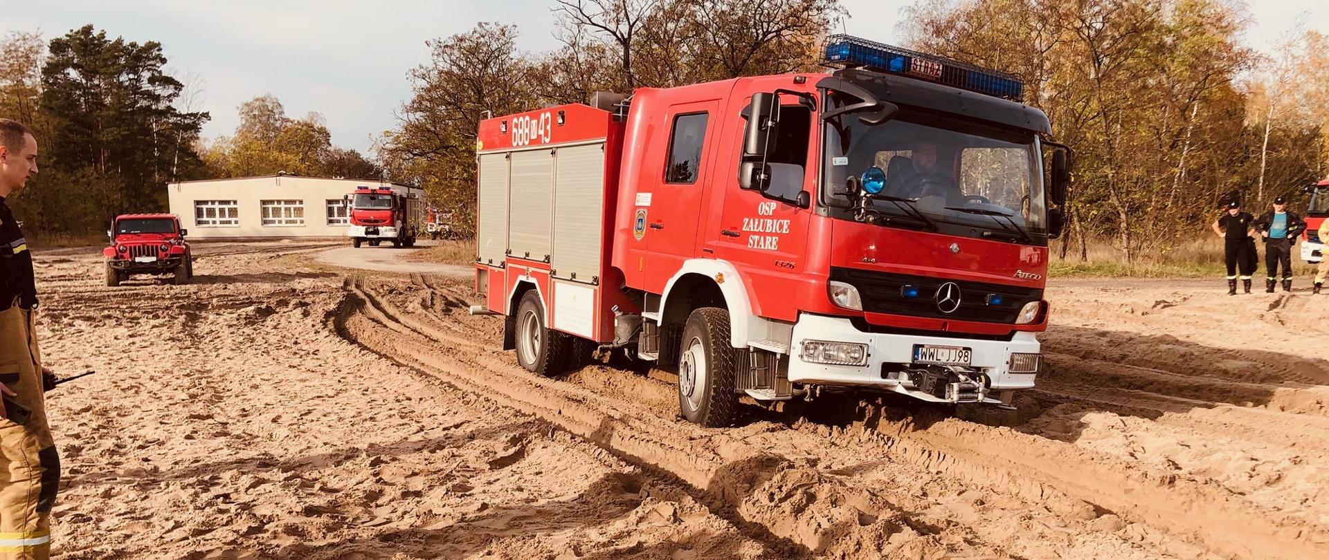 Na pierwszym planie widać czerwony samochód pożarniczy jadący po piaskowej drodze. Auto jest przechylone na lewą stronę. Po bokach widoczni są strażacy obserwujący ruch auta. W tle widać inne pojazdy oraz szary budynek.