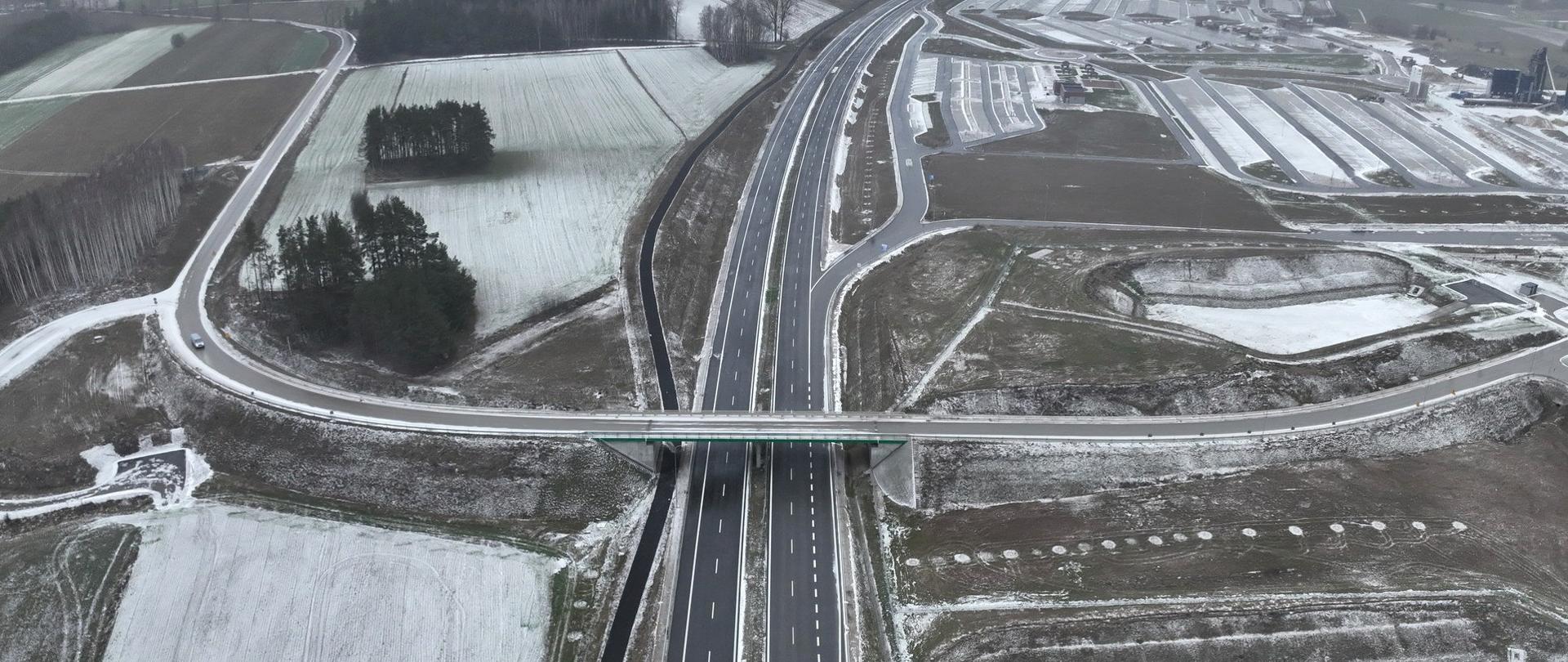 Widok z lotu ptaka na dwujezdniową drogę ekspresową z wiaduktem nad jezdnią, otoczoną polami częściowo pokrytymi śniegiem. Krajobraz zimowy, z szarym, pochmurnym niebem w tle i rozległymi terenami rolniczymi po obu stronach drogi.