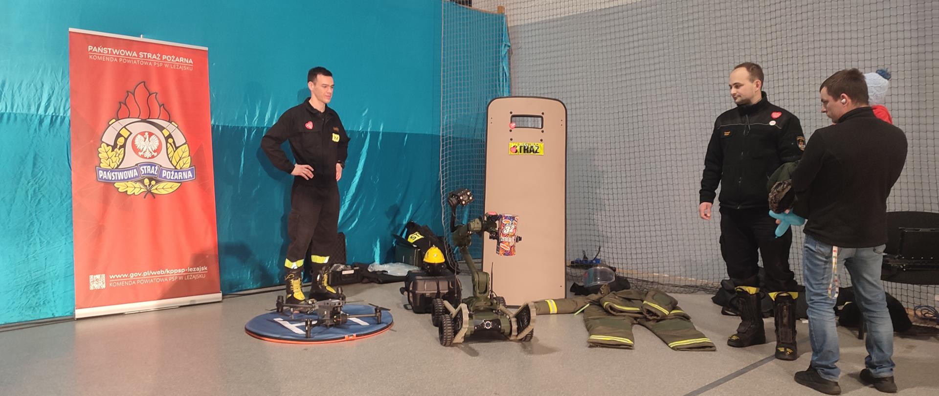Na zdjęciu widzimy strażaków prezentujących sprzęt, między innymi robota, tarcze balistyczną. W tle widzimy baner Państwowej Straży Pożarnej.