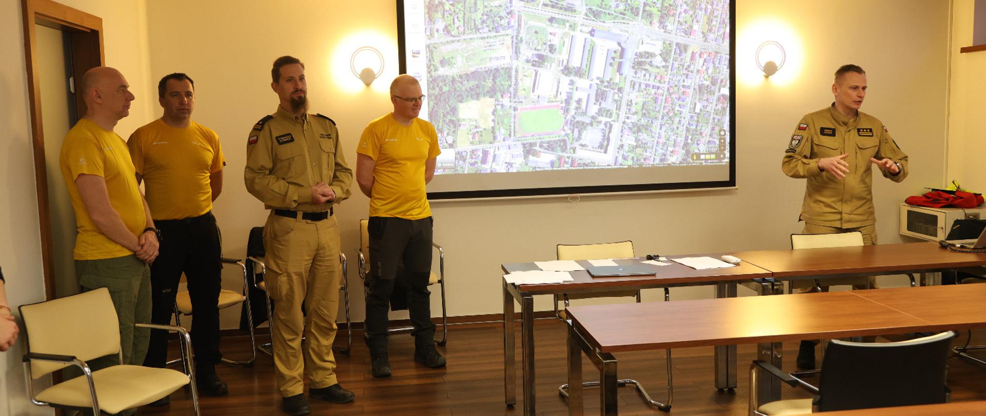 Na zdjęciu widać grupę mężczyzn w 2 w mundurach strażackich koloru piaskowego z naszywkami flagi Polski oraz emblematami jednostki PSP oraz trzech mężczyzn w żółtych koszulkach instruktorskich. Zdjęcie zrobione w nowoczesnej sali wykładowej z stołami ustawionymi w rzędach, w tle ekran projekcyjny z wyświetloną mapą rejonu. 