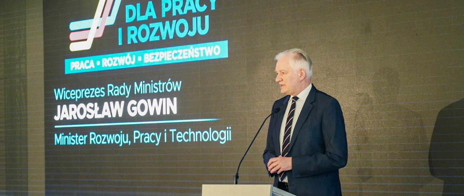 Na pierwszym planie stoi Premier Jarosław Gowin, w tle ścianka wizyjna z logo Plan dla Pracy i Rozwoju