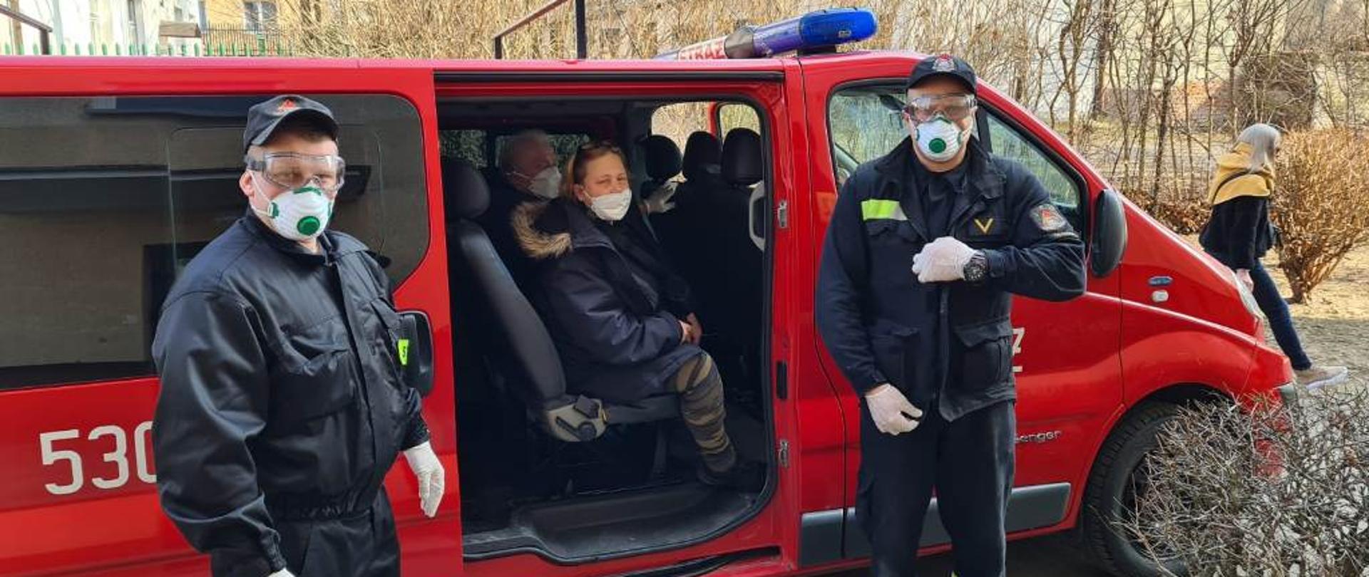 Dwie osoby siedzą w samochodzie strażackim typu bus, przy pojeździe stoją dwaj strażacy zabezpieczenie w maseczki typu ffp3, okulary i rękawiczki lateksowe
