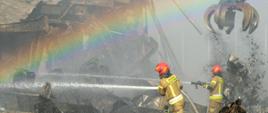 Dwóch strażaków PSP w ubraniach specjalnych i hełmach gasi pożar hałdy odpadów za pomocą prądów wody; w tle widoczny dźwig z chwytakiem oraz tęcza powstała w rozproszonej wodzie.