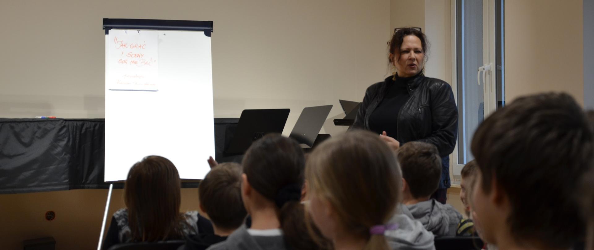 Sala wykładowa, wykładowca prowadzi warsztaty dla dzieci, na scenie biała tablica z rozrysowanymi schematami, grupa uczniów siedzi na widowni