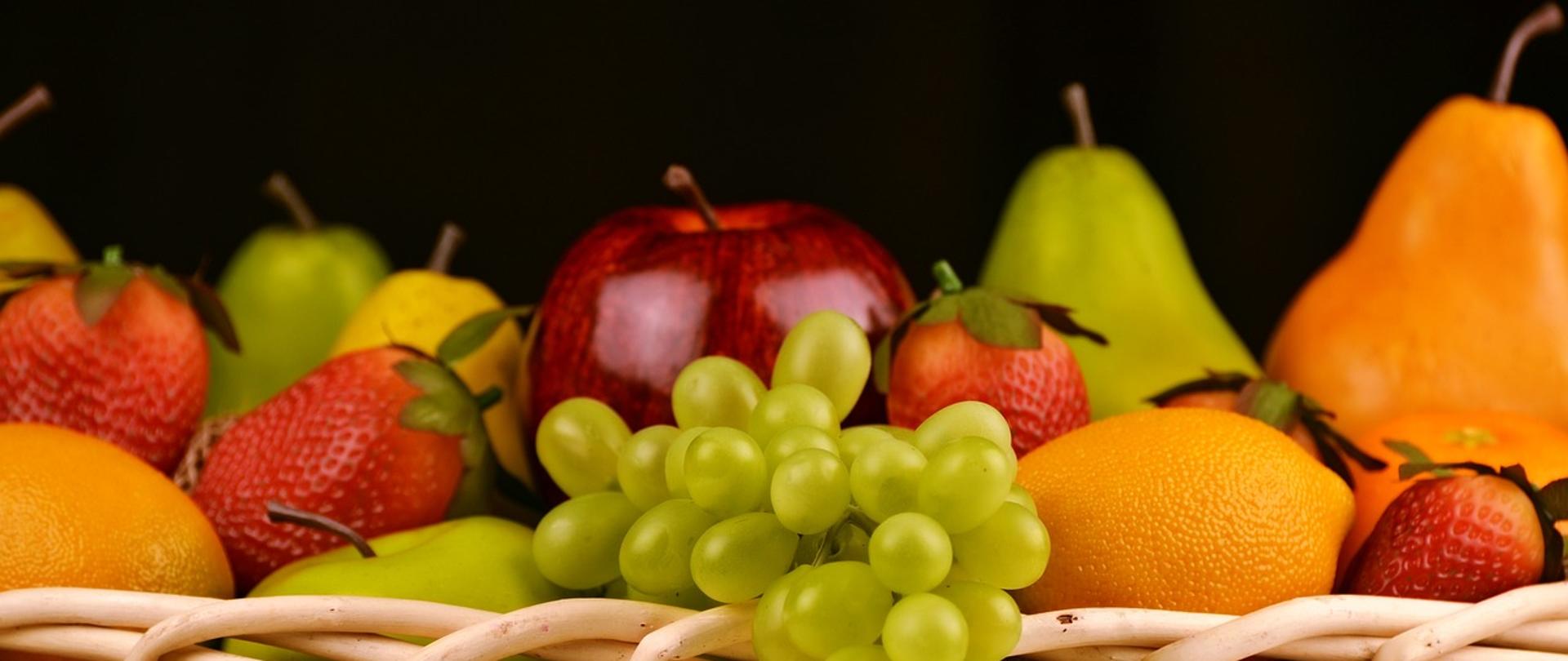 zdjęcie owoców w koszu wiklinowym, na pierwszym planie kiść zielonego winogrona, pomarańcza, cztery truskawki, czerwone jabłko, na drugim planie dwie gruszki w kolorze pomarańczowym oraz zielonym