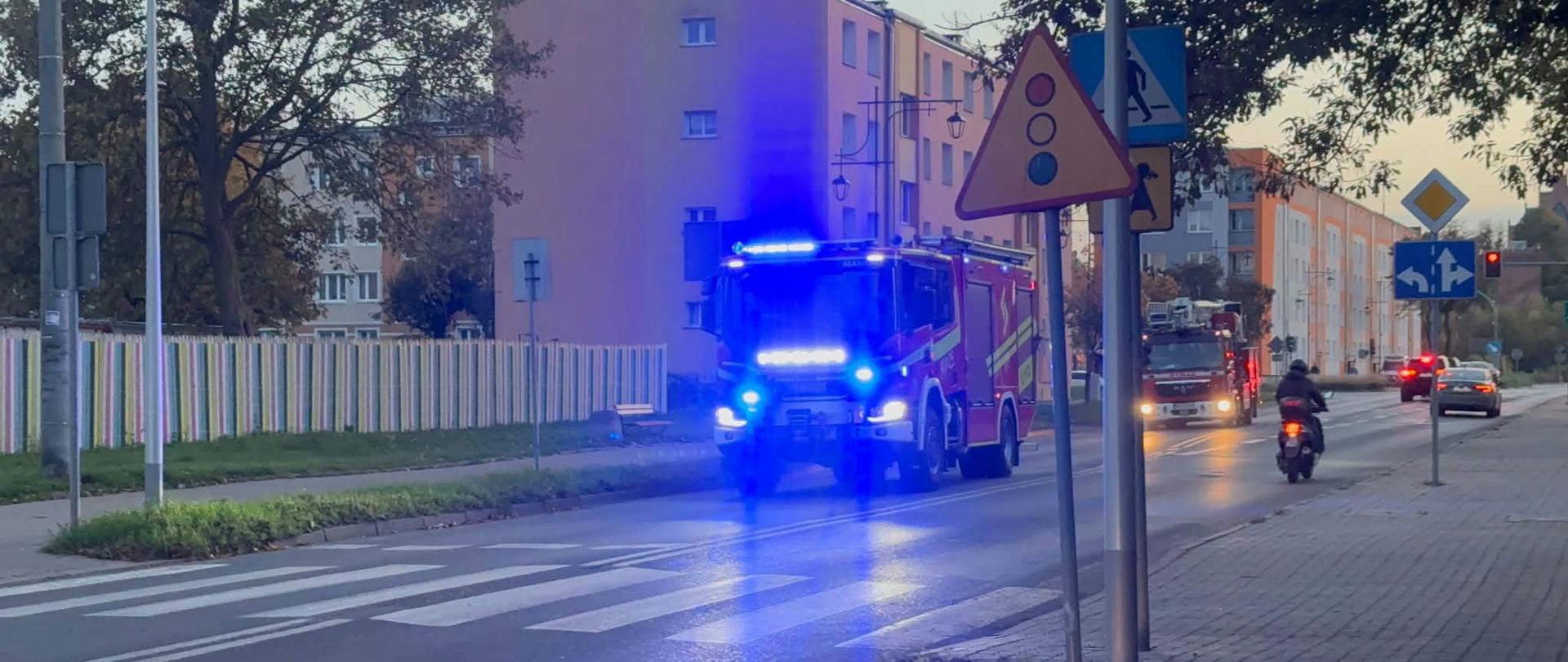 Ulicami miasta jadą wa duże samochody strażackie. Widać poświatę niebieskiego światła.