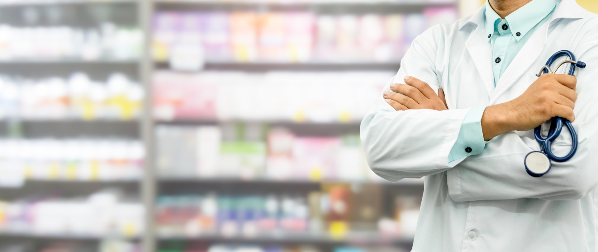 mężczyzna w białym fartuchu trzyma stetoskop, w tyle regały wypełnione lekami