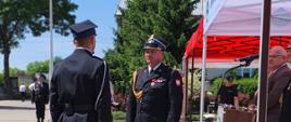 Na zdjęciu przedstawiono moment przyjęcia meldunku podczas uroczystego apelu z okazji Gminnych Obchodów Dnia Strażaka i jubileuszu 100-lecia OSP w Brańszczyku. W centralnej części kadru naprzeciw siebie stoją dwaj funkcjonariusze Państwowej Straży Pożarnej ubrani w czarne mundury wyjściowe z czapkami. Po prawej stronie widoczna jest mównica oraz fragment namiotu przygotowanego dla uczestników uroczystości. W tle znajdują się strażacy, zaproszeni goście oraz drzewa otaczające plac. Pogoda jest słoneczna, a niebo bezchmurne.