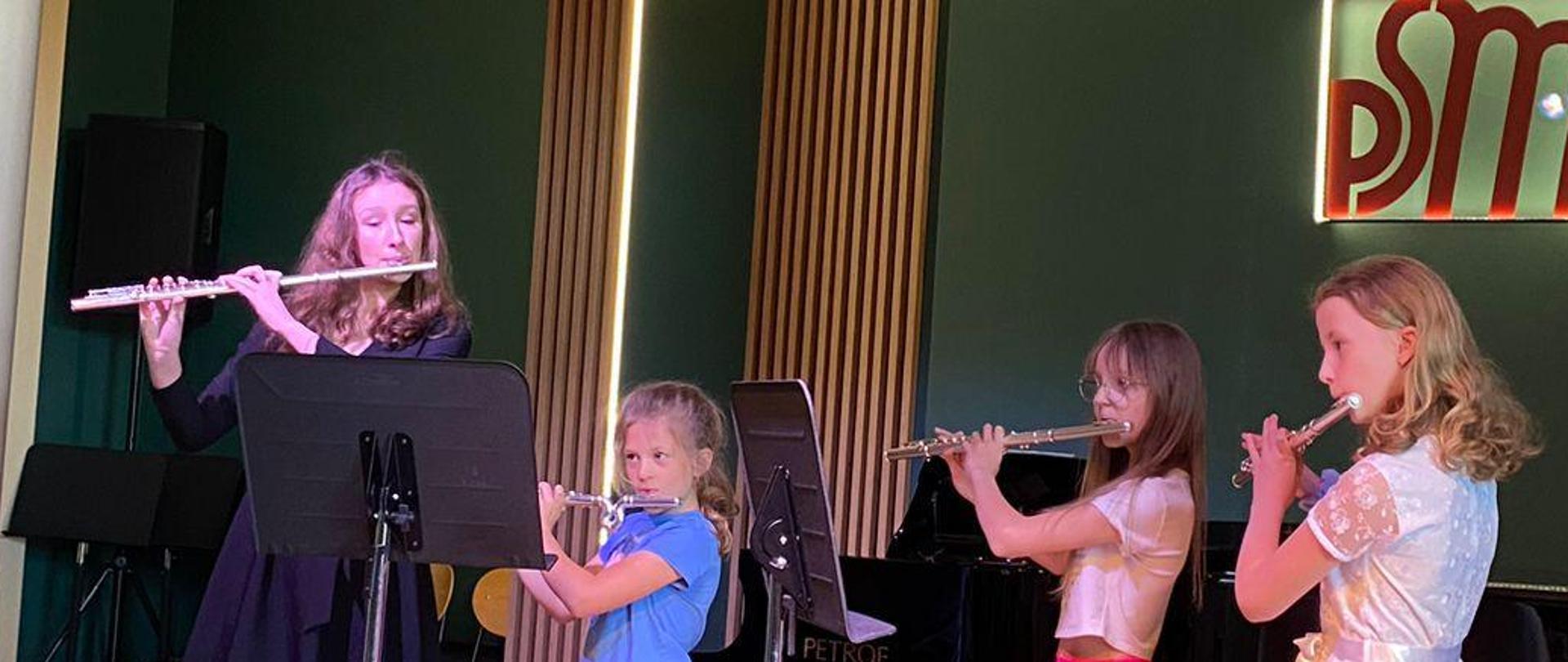 4 uczennice grające na fletach w tle widoczny jest napis z logo szkoły oraz ściana w kolorze zielonym z drewnianymi lamelami