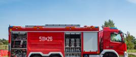 Duży czerwony pojazd gaśniczy na tle niebieskiego nieba. Obok widoczny zbiornik wodny. Pojazd jest przedstawiony z boku, widać numer operacyjny pojazdu 511-26 . Samochód ma otwarte skrytki boczne, widoczny jest sprzęt pożarniczy, armatura wodno pianowa