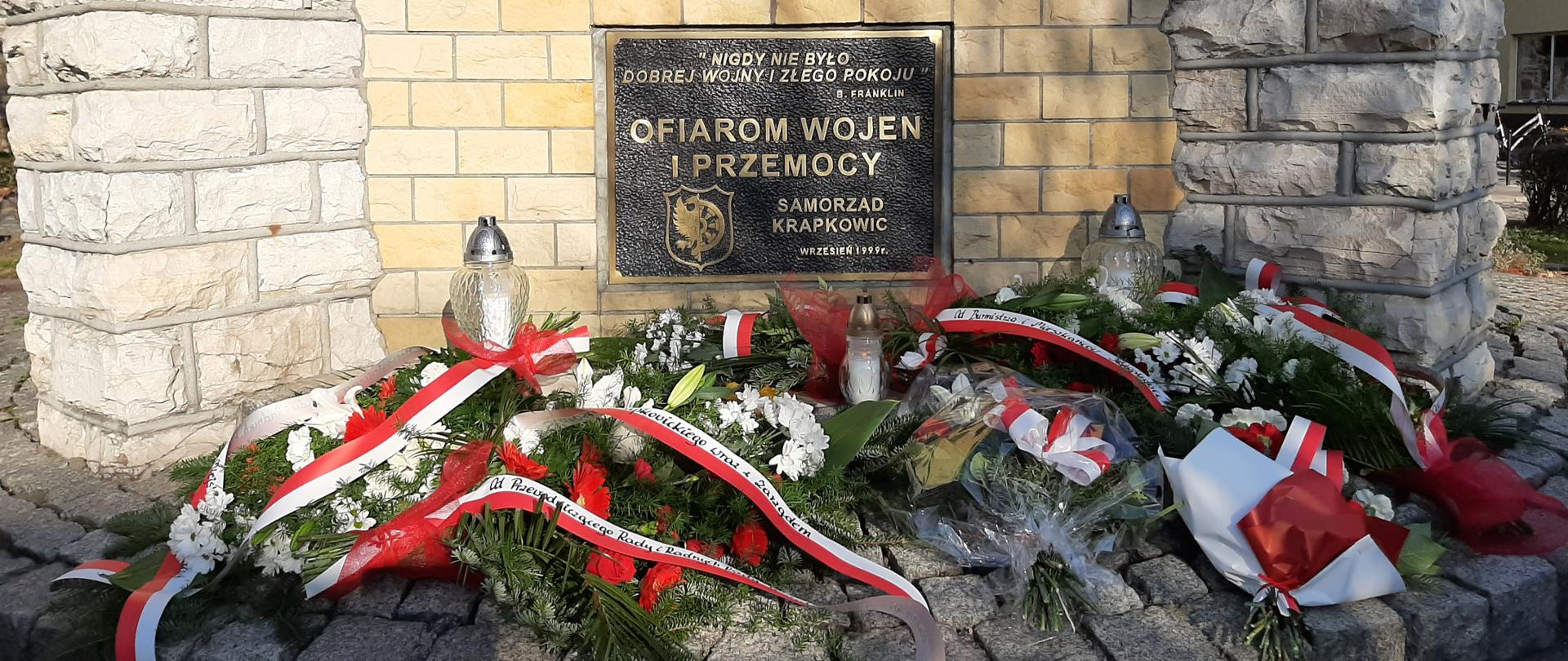 Zdjęcie przedstawia cokół pomnika Ofiar Wojen i Przemocy. Na cokole pamiątkowa tablica z cytatem B.Franklina: "Nigdy nie było dobrej wojny i złego pokoju", poniżej napis "OFIAROM WOJEN I PRZEMOCY", a jeszcze niżej herb Krapkowic oraz informacja o fundatorze tablicy "SAMORZĄD KRAPKOWIC" oraz data Wrzesień 1999r. U podnóża pomnika złożone wiece i wiązanki kwiatów z biało-czerwonymi wstęgami.