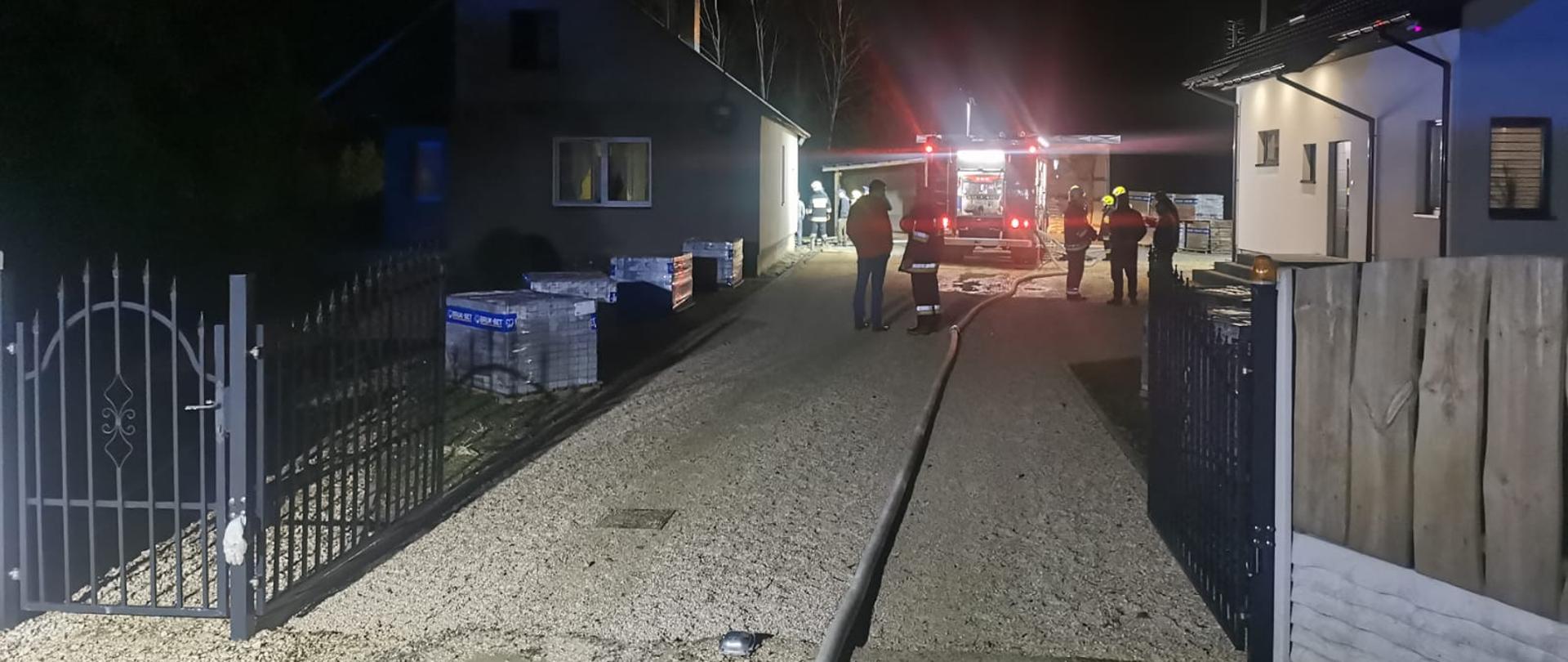 Pora nocna, prywatna posesja widziana z drogi, na pierwszym planie ogrodzenie w oddali samochód strażacki do którego podłączony jest wąż pożarniczy oraz po obu stronach budynki