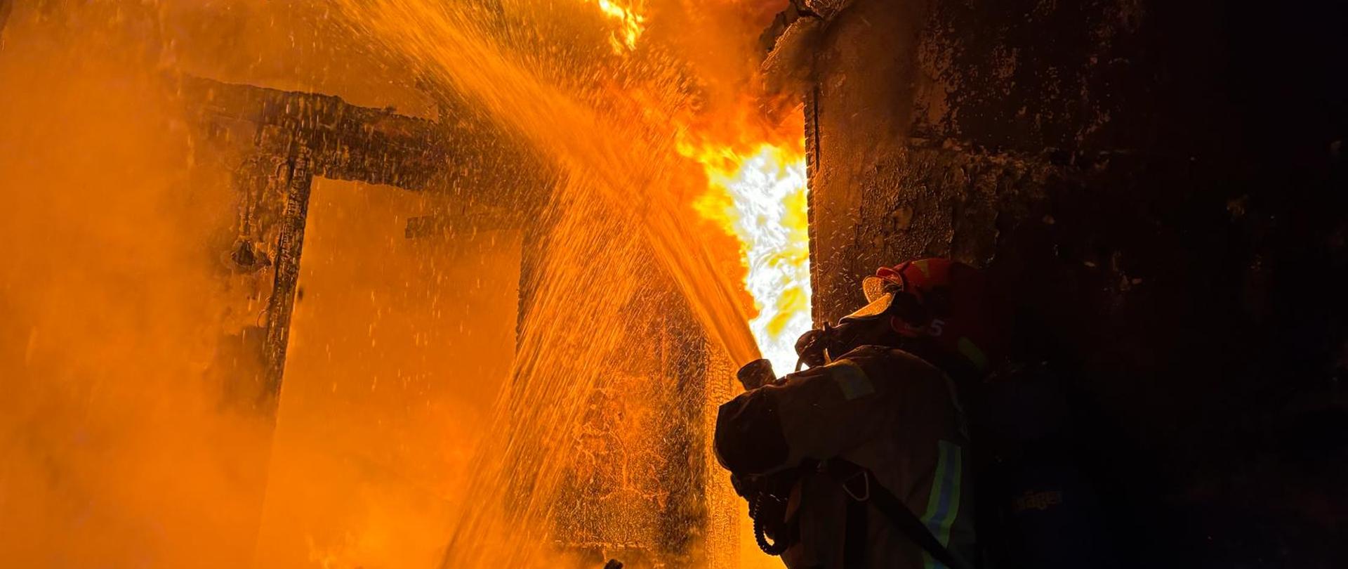 Strażak w hełmie na poddaszu podaje prąd wody na palący się drewniany strop.