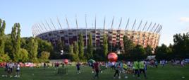 Na zielonej trawie odbywa się piknik, wszędzie są ludzie, w tle drzewa i Stadion Narodowy.