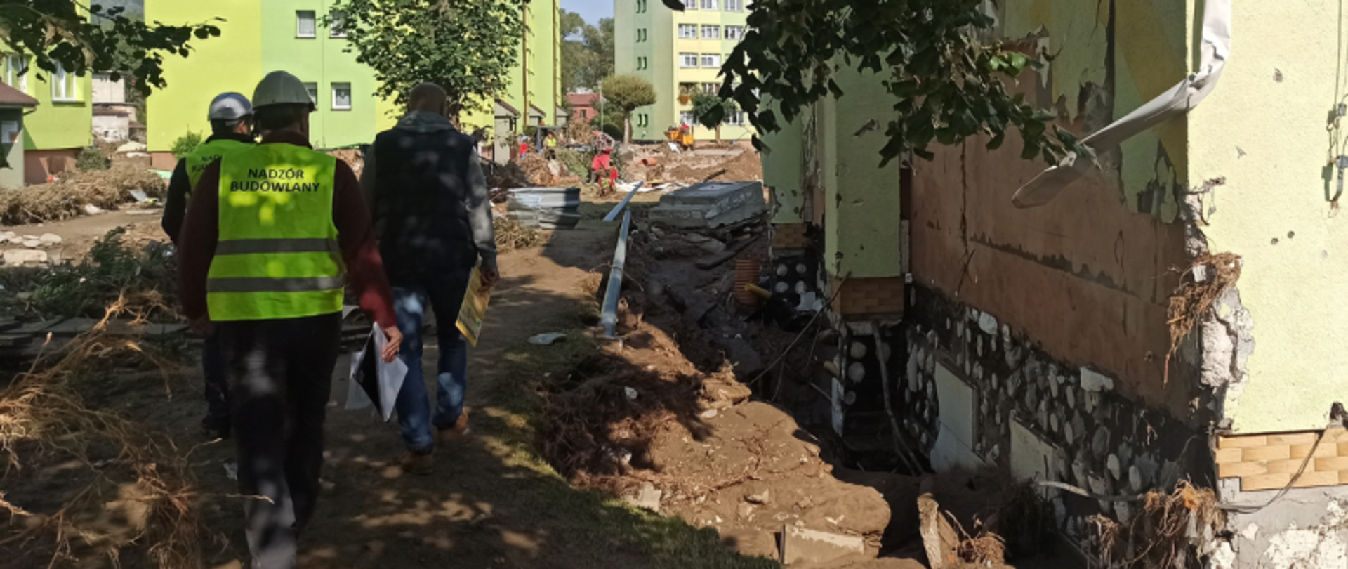 Plac budowy z osobami i nadzorem budowlanym; wokół gruz, materiały i zielone budynki w tle.