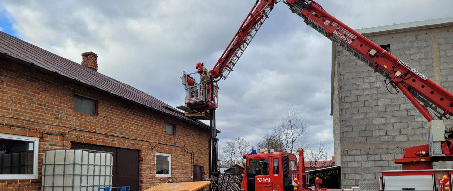 Podnośnik strażacki stoi na podwórku posesji, na której znajdują się dwa budynki. Na jednym z nich został uszkodzony dach w wyniku silnego wiatru. Podnośnik ma rozłożoną drabinę z koszem, w którym znajduje się dwóch strażaków, którzy naprawiają dach na budynku.