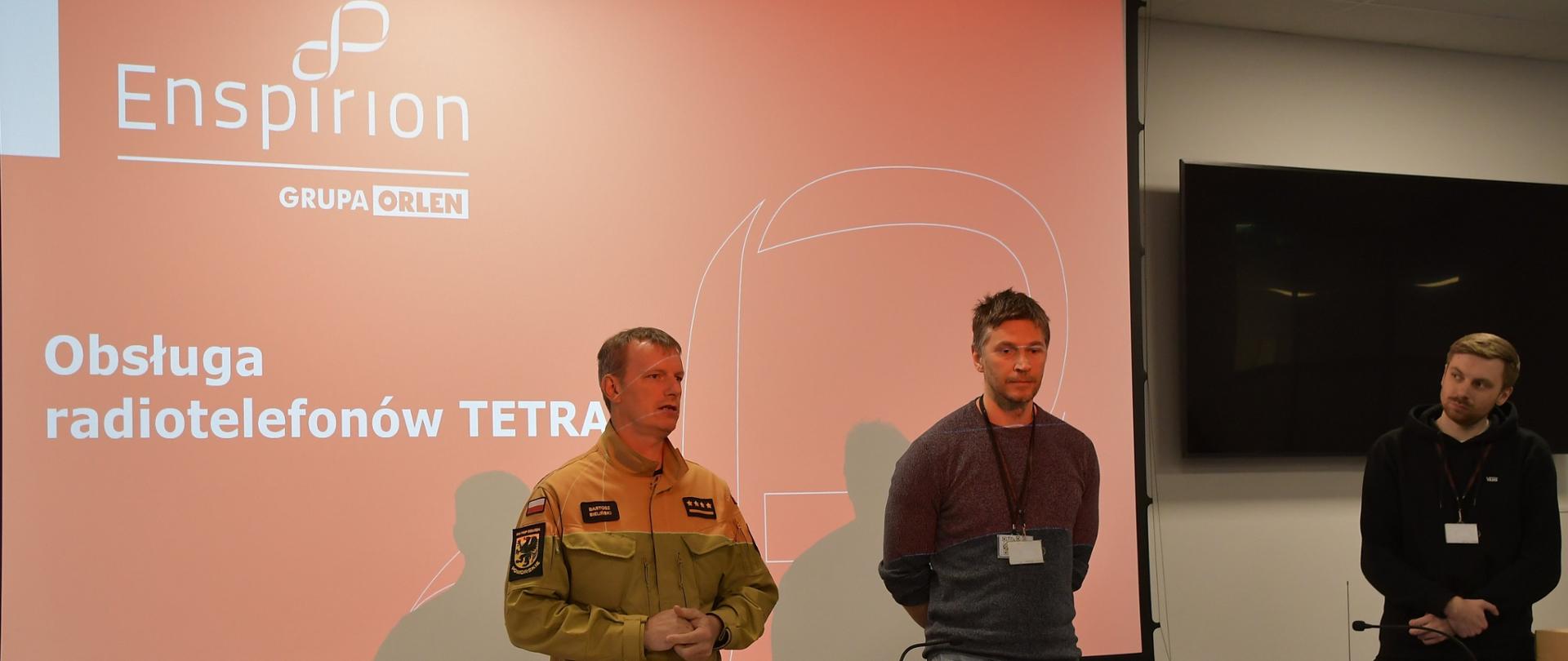 Strażak w umundurowaniu koloru musztardowego stoi obok dwóch mężczyzn na stole leżą komputery na ekranie jest wyświetlane logo i napis Grupa Orlen obsługa radiotelefonów Tetra.