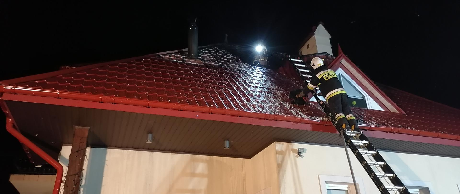 Na zdjęciu widać budynek jednorodzinny z uszkodzonym w wyniku pożaru dachem pokrytym blachodachówką. Do ściany budynku przystawiona jest drabina pożarnicza, na której pracuje strażak OSP, ubrany w ubranie specjalne. Z dziury na dachu widać strażaka OSP w umundurowaniu specjalnym.