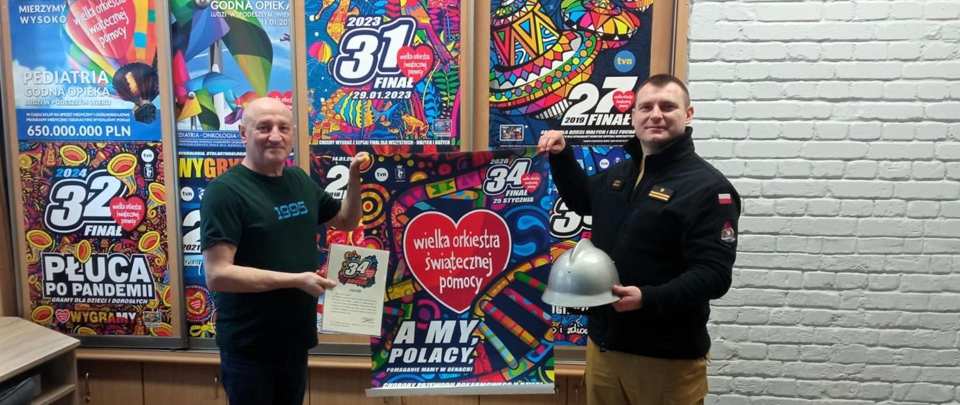 Zdjęcie przedstawia moment przekazania przez Zastępcę Komendanta Powiatowego Państwowej Straży Pożarnej w Gołdapi mł. bryg. Michała Herman przedstawicielowi sztabu Wielkiej Orkiestry Świątecznej Pomocy w Gołdapi egzemplarz hełmu strażackiego pochodzący z listopada 1971 roku oraz voucher umożliwiający darczyńcom wizytę w siedzibie gołdapskich strażaków i uczestnictwo w „Dniu z życia Strażaka”, które zostaną zlicytowane podczas 34. Finału WOŚP