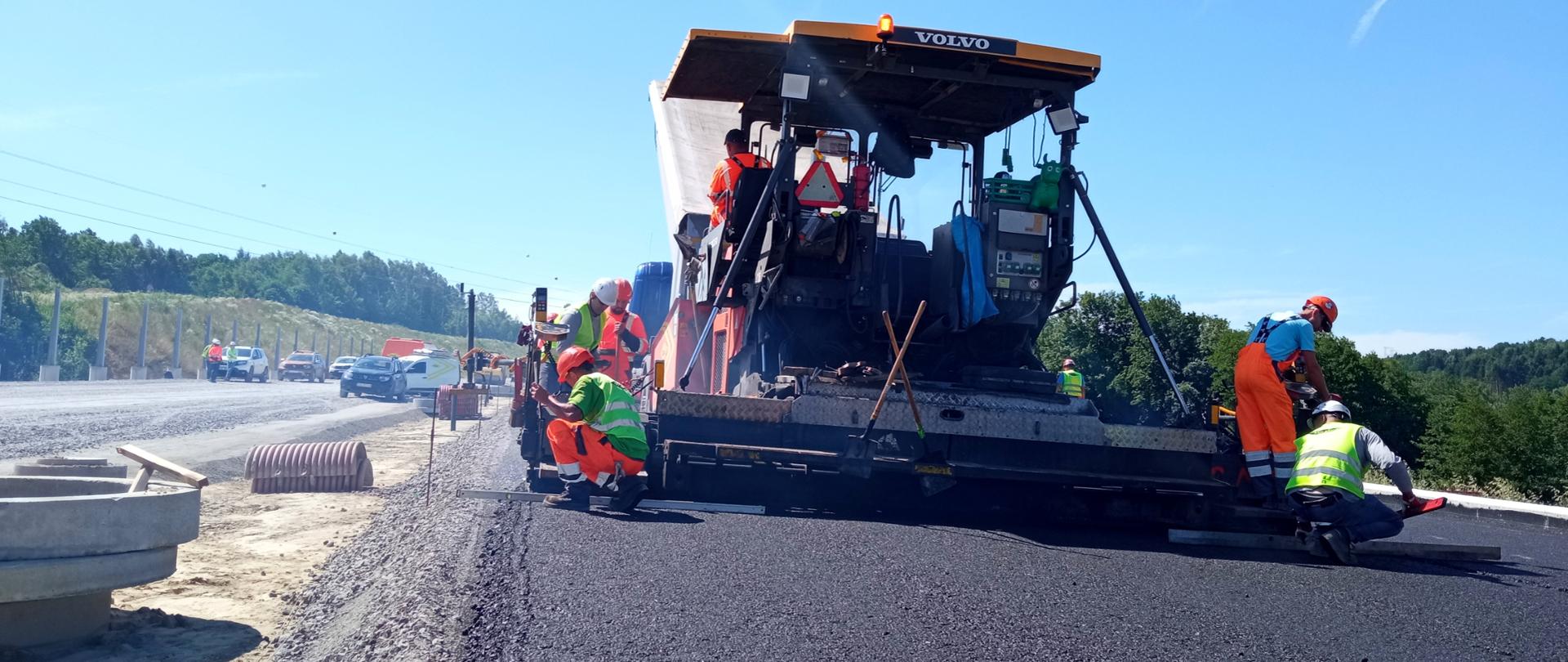 Świeci słońce. Widok z poziomu ziemi na układanie masy. Widać pracujące rozściełącze oraz robotników. 