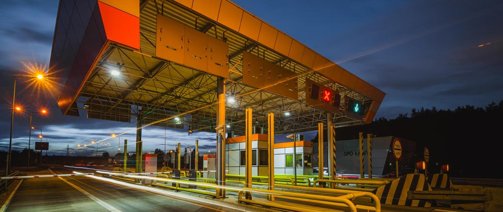 Miejsce Poboru Opłat na autostradzie A4 w porze wieczornej, z charakterystycznym zadaszeniem nad pasami ruchu, sygnalizacją świetlną nad bramkami oraz śladami świateł przejeżdżających pojazdów na tle ciemniejącego nieba.