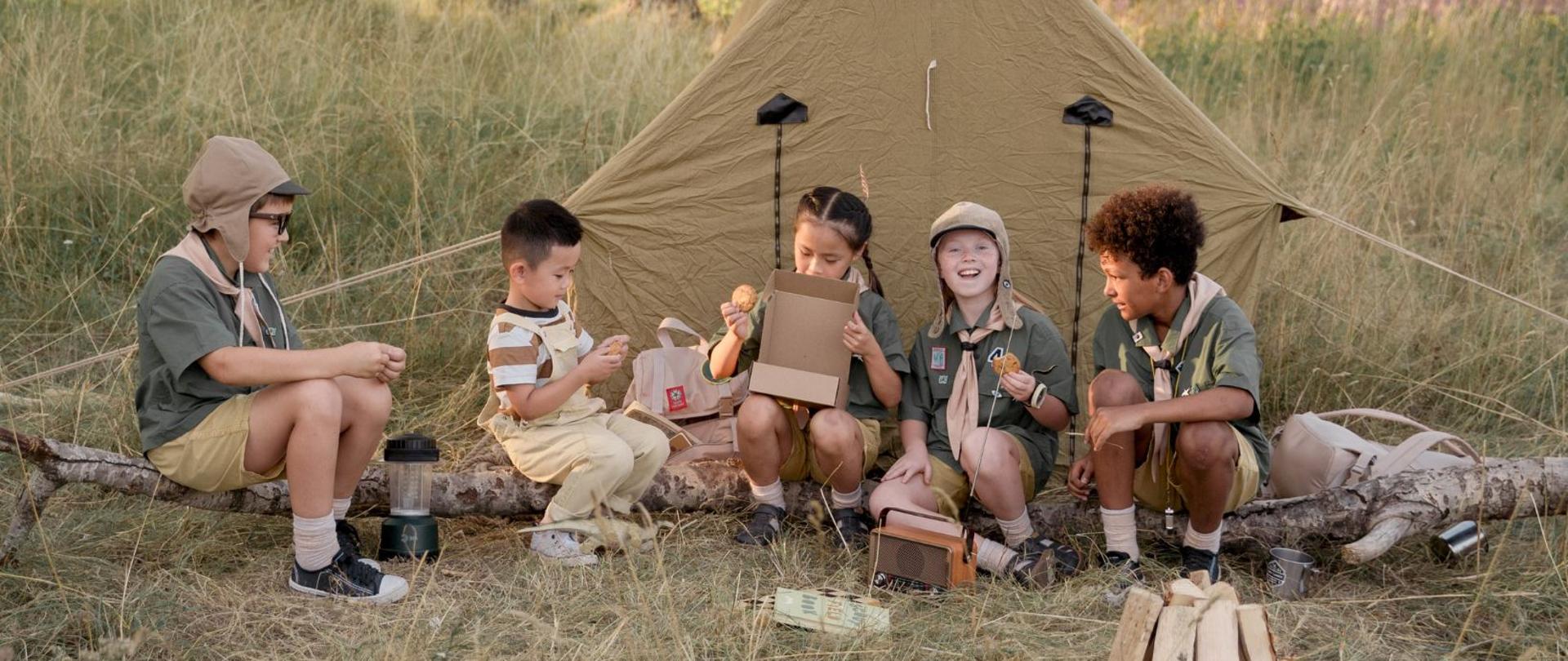 Dzieci siedzące przed namiotem przygotowują się do rozpalenia ogniska.