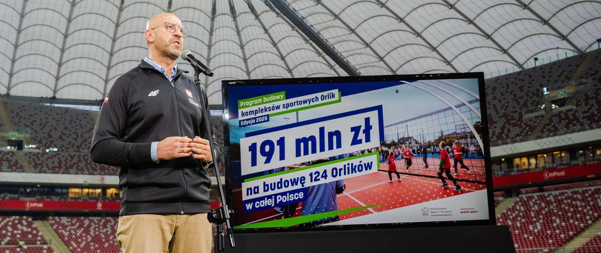Minister of Sport and Tourism speaks to the microphone at the PGE Narodowy national stadium. Screen with basic information concerning the programme devoted to the construction of Orlik pitches.