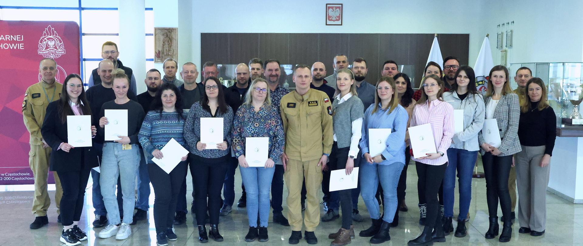 Pamiątkowe zdjęcie grupowe uczestników szkolenia inspektorów ochrony przeciwpożarowej (26 osób) w towarzystwie Zastępcy Komendanta Centralnej Szkoły PSP, kobiety oraz dwóch funkcjonariuszy CS PSP
