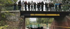 Na zdjęciu znajduje się śluza w Pakości. Składa się z ceglanych ścian, kamiennych umocnień, metalowych barierek. W komorze śluzy jest woda. Nad śluzą mostek. Na mostku stoi młodzież szkolna. Machają w stronę fotografa. 