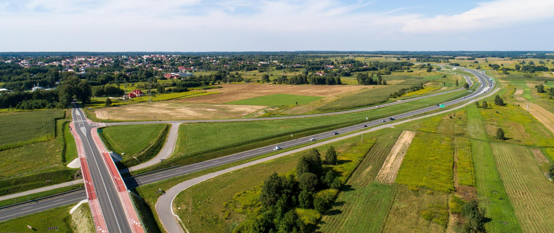 Widok z góry na odcinek drogi ekspresowej S19 Sokołów Młp. - Stobierna. Lato, świeci słońce, zieleń dookoła drogi. Nad trasą ekspresówki przebiega wiadukt.. W tle widać zabudowania.