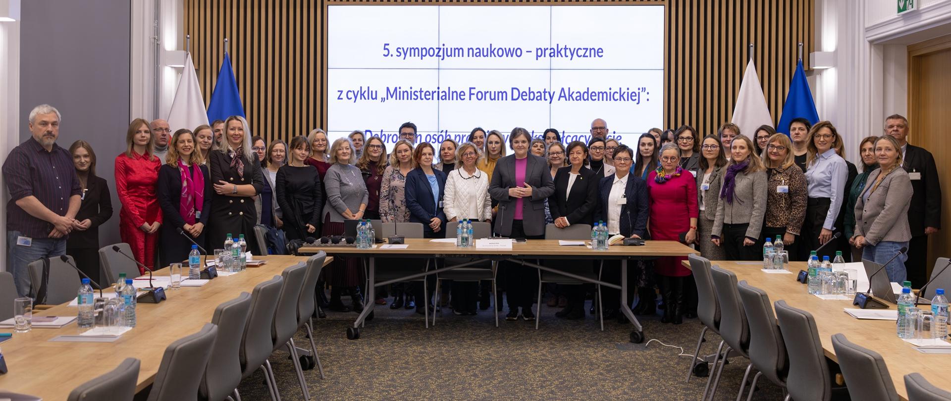 Zdjęcie zbiorowe, pod wbudowanym w ścianę z cienkich pionowych listewek wielkim ekranem z napisem 5. sympozjum naukowo-praktyczne z cyklu Ministerialne Forum Debaty Akademickiej stoi grupa kilkudziesięciu osób, pośrodku wiceminister Zioło-Pużuk.