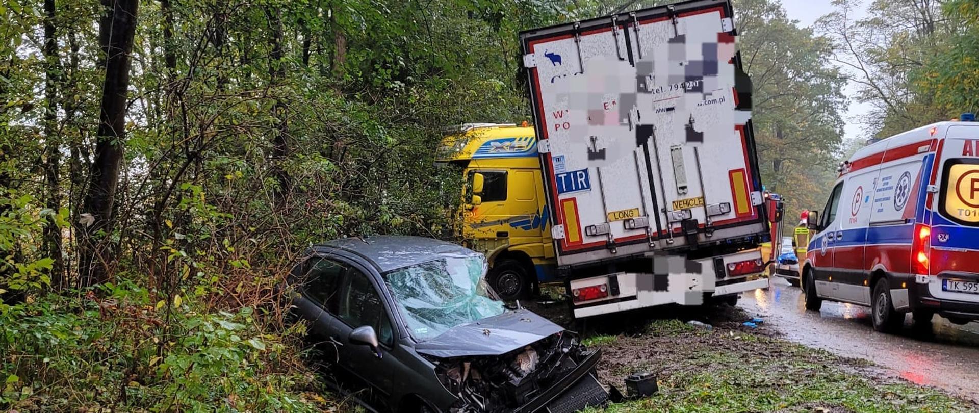 Na 1 planie pozostałości karoserii z pojazdów, za nimi po lewej stronie w rowie ze zgniecionym przodem samochód osobowy, za nim stojąca tyłem naczepa a za nią widać przód ciągnika siodłowego, po prawej stronie stoi pojazd karetki pogotowie a przed nią strażak PSP