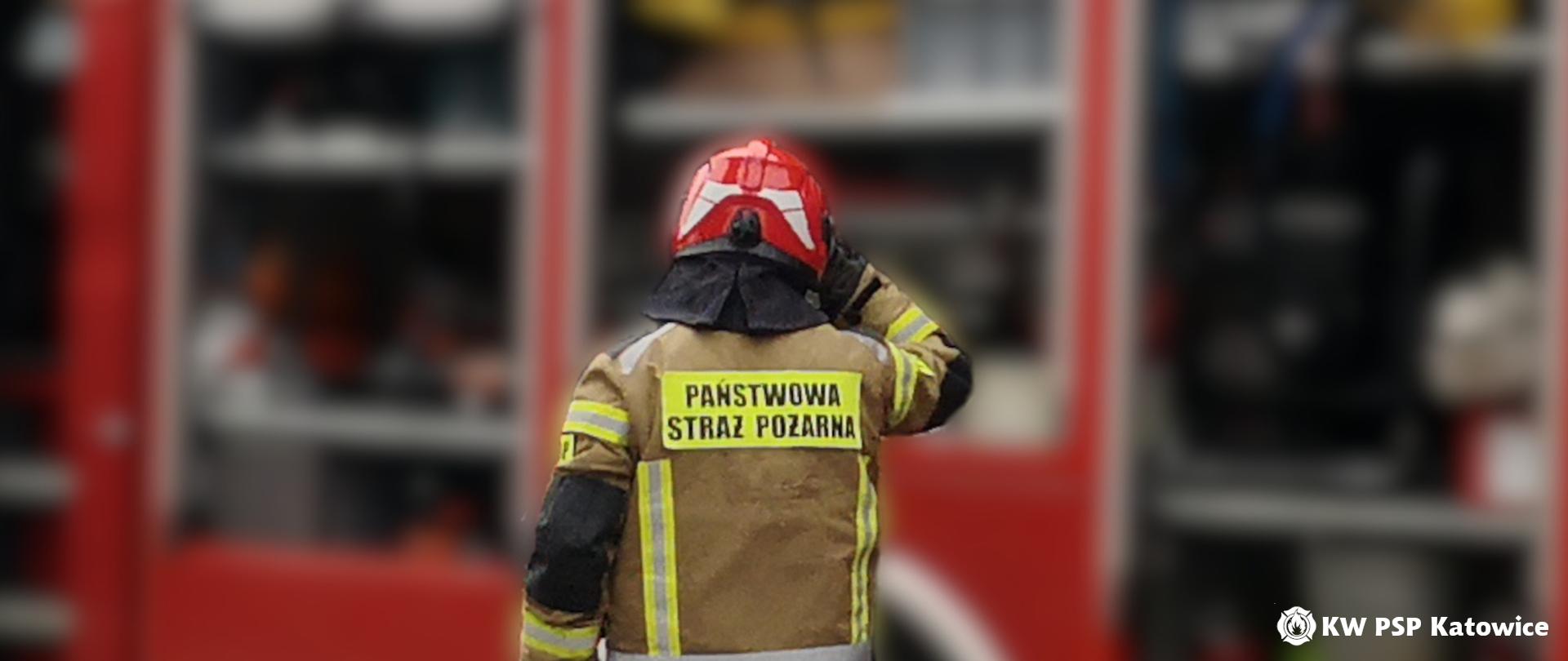 Strażak w ubraniu specjalnym i hełmie stoi tyłem na tle czerwonego samochodu pożarniczego z otwartymi skrytkami
