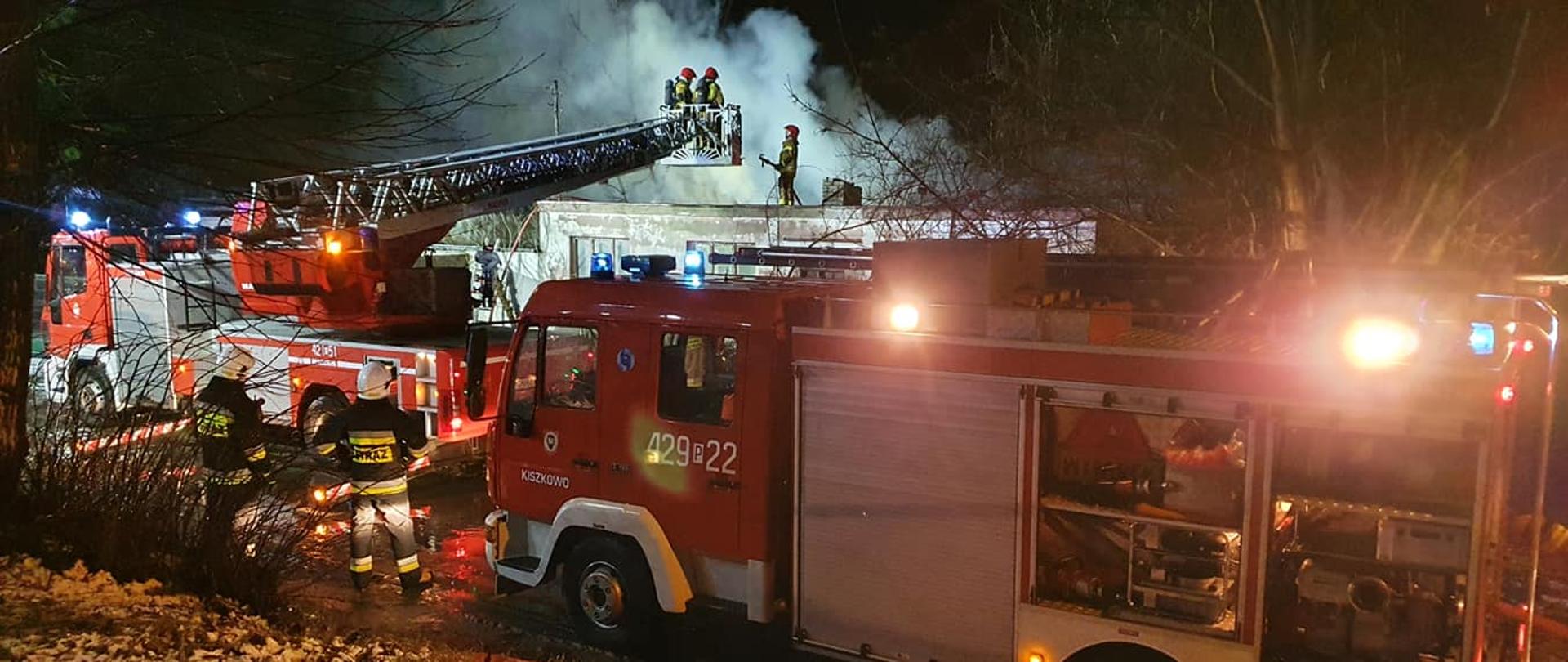Na zdjęciu widać samochody pożarnicze biorące udział w akcji. W koszu autodrabiny, który jest bezpośrednio nad dachem budynku znajdują się dwóch strażaków. Jest pora nocna, nad budynkiem unosi się dym. Na dachu stoi strażak z wężem pożarniczym