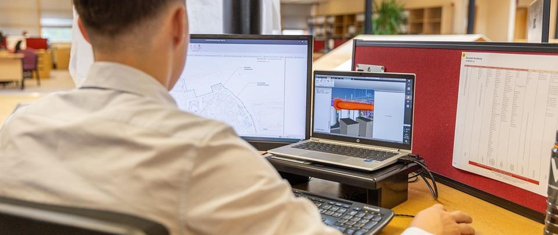 Mężczyzna podczas pracy na stanowisku wyposażonym w laptopa, dwa monitory ekranowe oraz klawiaturę i mysz w pomieszczeniu biurowym