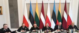 Mężczyźni w mundurach siedzący i prowadzący rozmowę przy stole. Przed każdym z nich na stole stoi wizytówka. W tle flagi Polski, Estonii, Litwy oraz Łotwy