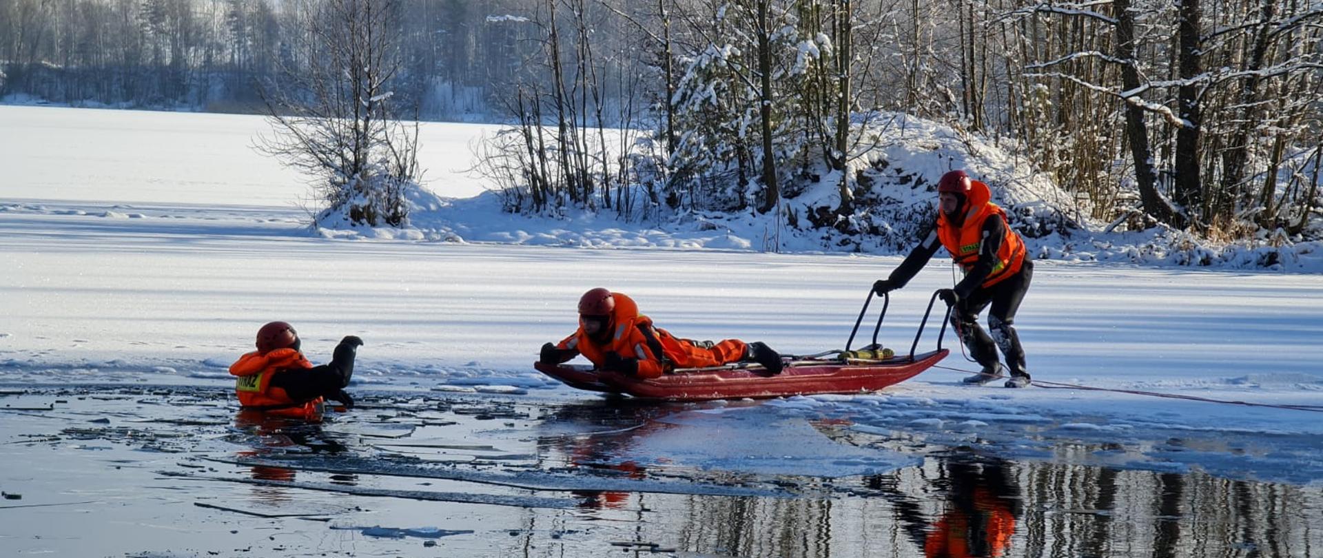 Zamarznięta tafla zbiornika wodnego, widoczna popękana powierzchnia - unoszą się kry. W środku kadru widoczne sanie lodowe ratunkowe na których leży ratownik, funkcjonariusz straży pożarnej drugi funkcjonariusz asekuruje za uchwyty sanie lodowe trzeci z funkcjonariuszy zanurzony w wodzie do połowy jest ewakuowany z wody. W tle widoczne drzewa. Ćwiczenia przeprowadzane są około 10 m od linii brzegowej.