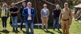 Na pierwszym planie, na trawiastym terenie przed dużym wojskowym namiotem, stoi grupa osób podczas oficjalnego spotkania. W centrum zdjęcia znajduje się Starosta w niebieskiej marynarce i dżinsach, który przemawia do zgromadzonych. Obok niego, po prawej stronie, stoi Komendant Powiatowy Państwowej Straży Pożarnej w piaskowym mundurze służbowym i czapce z daszkiem. W tle, za nimi, stoi kilka osób w strojach cywilnych oraz kamizelkach odblaskowych, przysłuchujących się wypowiedzi. Akcja dzieje się w słoneczny dzień, w warunkach polowych.