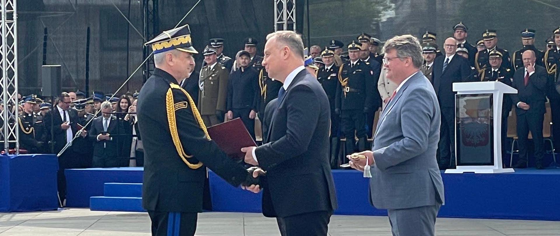 Pomorski komendant wojewódzki Państwowej Straży Pożarnej odbiera akt mianowania na wyższy stopień od prezydenta Rzeczypospolitej Polskiej. Obok stoi sekretarz stanu w Ministerstwie Spraw Wewnętrznych i Administracji z szablą. Przed nimi stoi trybuna honorowa z zaproszonymi gośćmi.