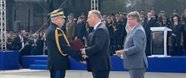 Pomorski komendant wojewódzki Państwowej Straży Pożarnej odbiera akt mianowania na wyższy stopień od prezydenta Rzeczypospolitej Polskiej. Obok stoi sekretarz stanu w Ministerstwie Spraw Wewnętrznych i Administracji z szablą. Przed nimi stoi trybuna honorowa z zaproszonymi gośćmi.
