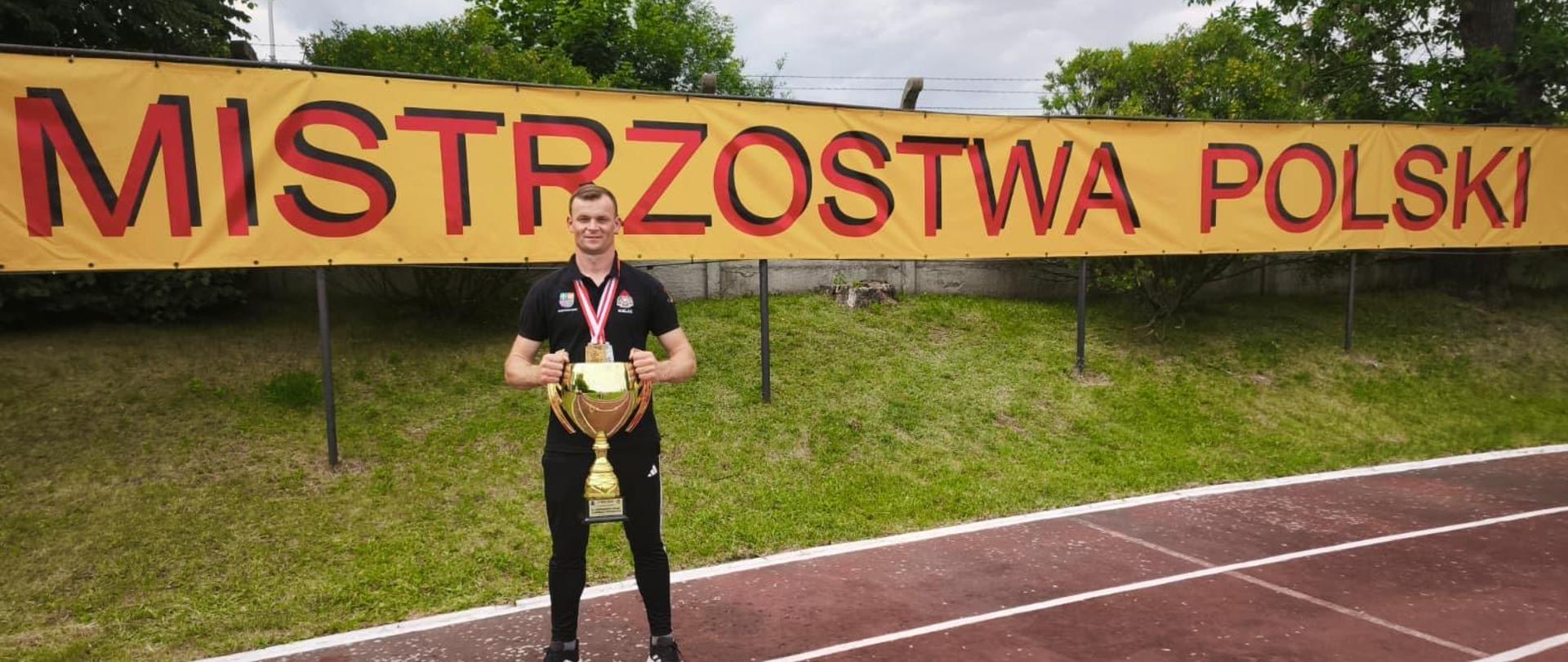Na zdjeciu widoczny jest kpt. Robert Zdonek trzymający puchar na tle baneru Mistrowstwa Polski, który na co dzień pełni funkcję Zastępcy Dowódcy zmiany.