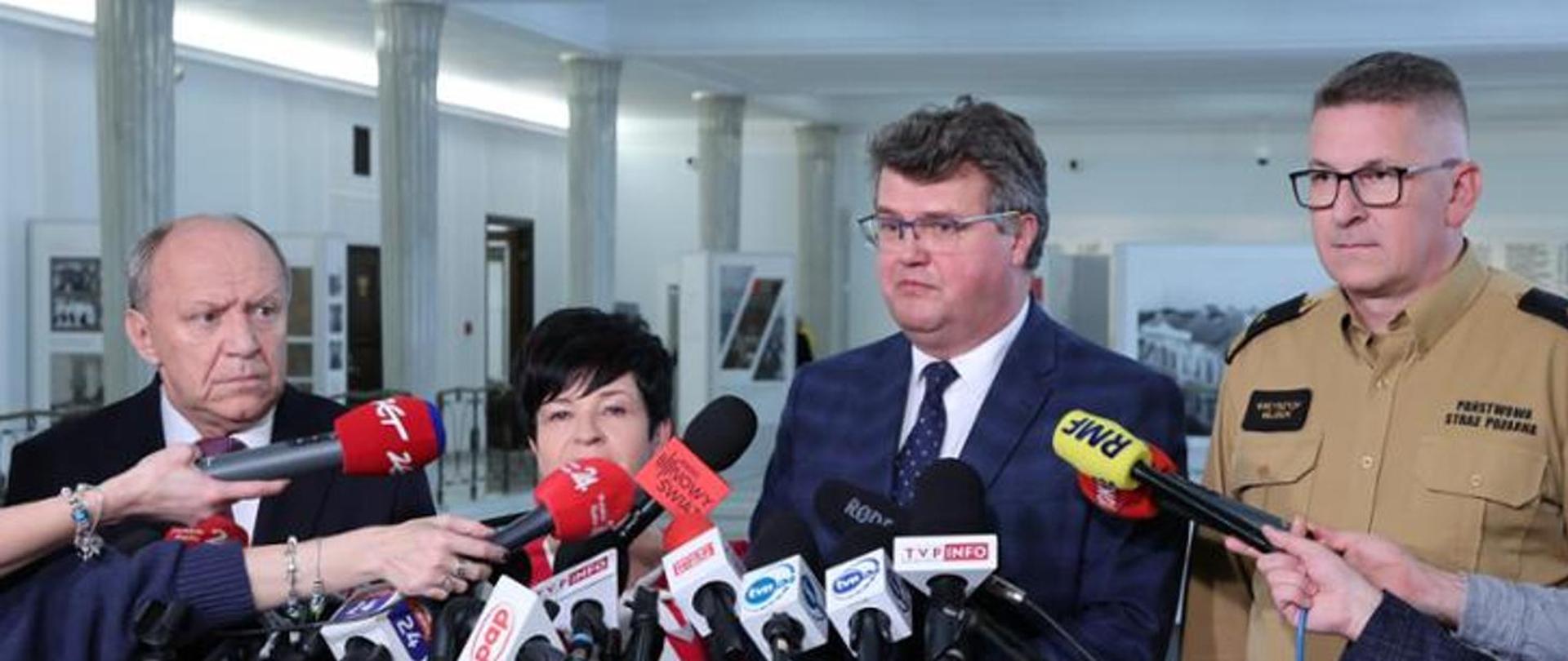 Na zdjęciu widzimy cztery osoby stojące przed mikrofonami podczas konferencji prasowej