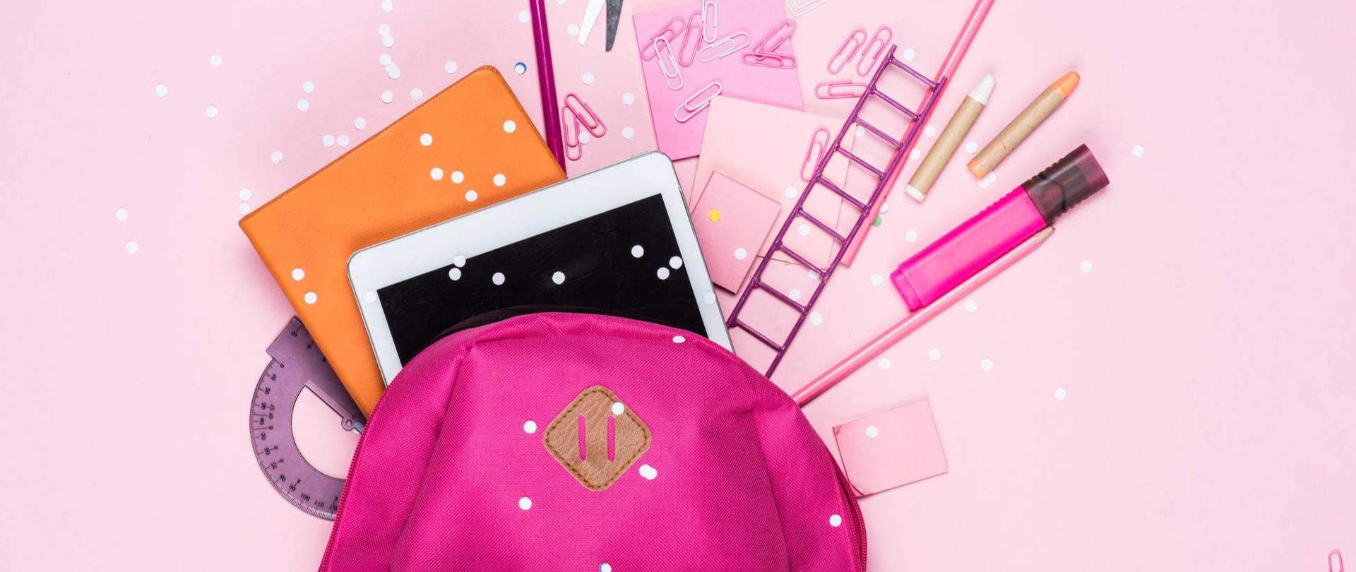 Na zdjęciu jest plecak i przybory szkolne takie jak na przykład: nożyczki, zeszyt, spinacze. Tło jest różowe.