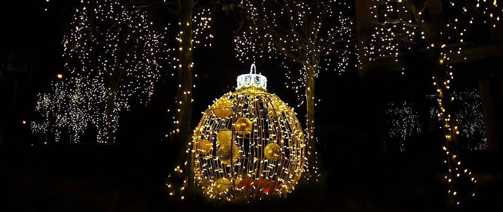 Zdjęcie nocne plenerowej instalacji 3d - bombka choinkowa oświetlona lampkami w tonacji żółtej, w tle drzewa także oświetlone lampkami choinkowymi.